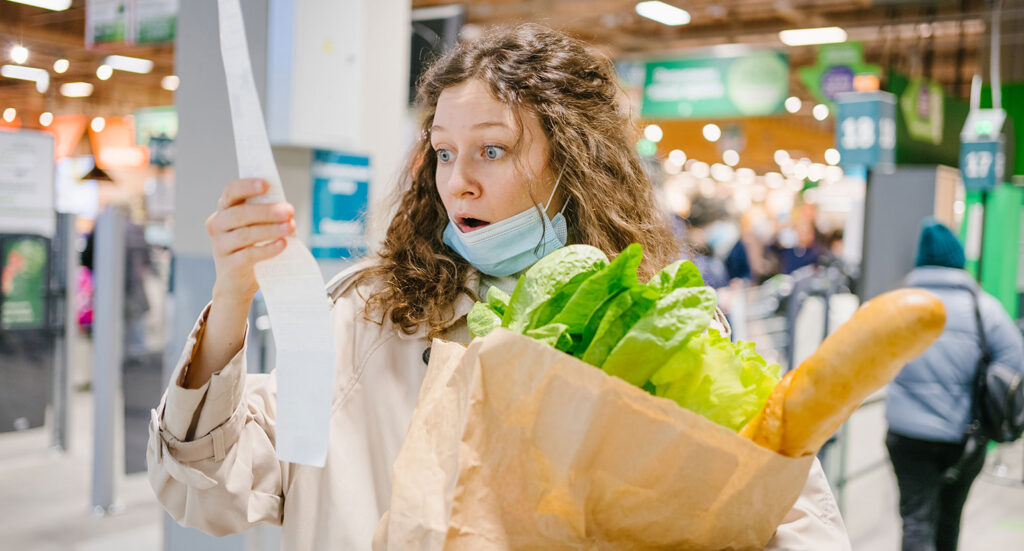 Woman with shocked expression looking at receipt with groceries in hand