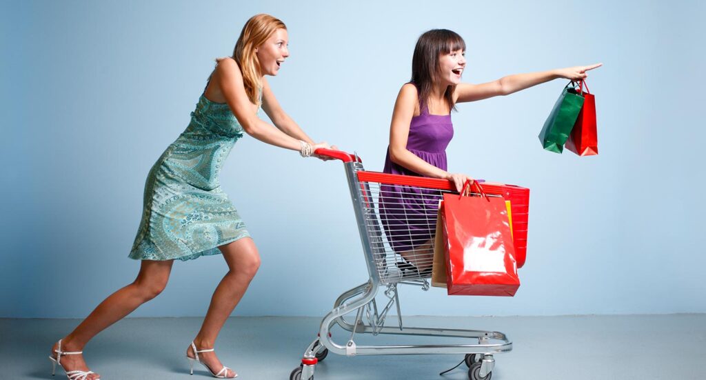 Woman pushing another woman holding shopping bags in shopping trolley
